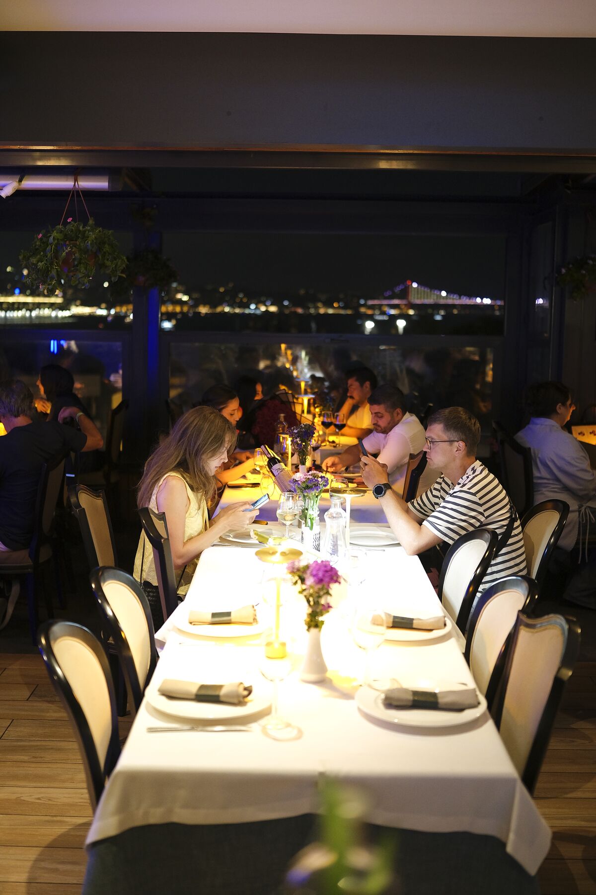 Guests dining at a candle-lit long table on the Pino Gare terrace at night, the illuminated Bosphorus Bridge through the glass